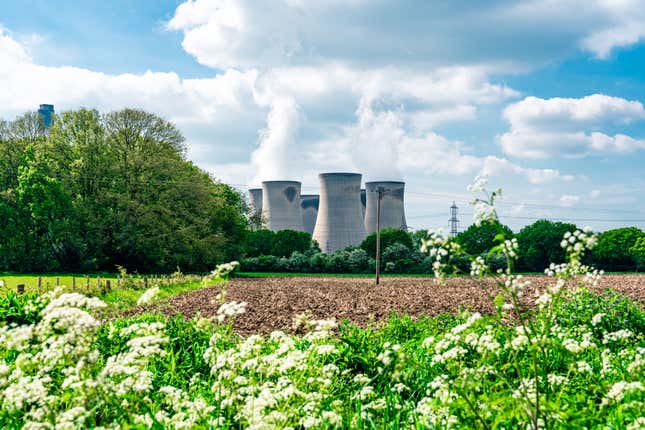 sunny day, power station in the background, with a field of green in the foreground, and green trees on the left