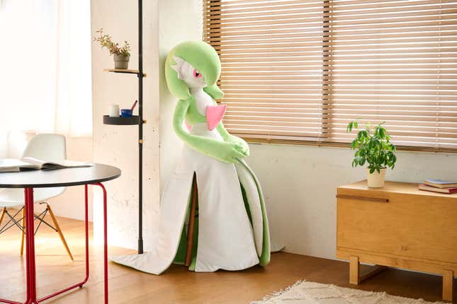 A Gardevoir life-sized plush sitting next to a window.