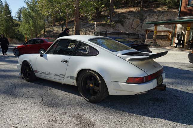 A white 1980s Porsche 911 with a big spoiler is parked near a red BMW M3