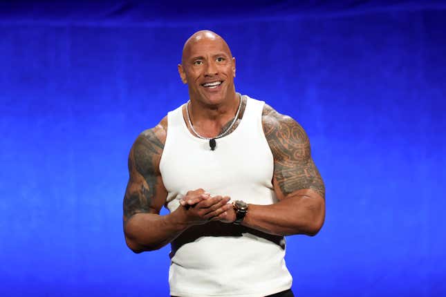 Dwayne Johnson wearing a white tank top with tattoos on both biceps, standing in front of a royal blue backdrop