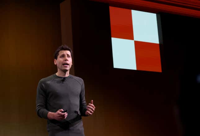 Sam Altman speaking on stage with a black and red square on the upper right corner of the backdrop behind him