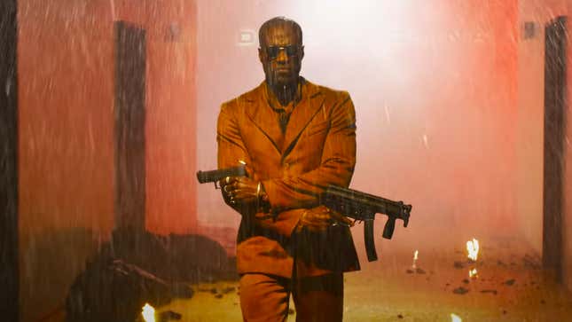 Yahya Abdul-Mateen II’s Morpheus dressed in an orange suit and wielding two guns with his arms crossing over one another in the midst of a fight in an office building.