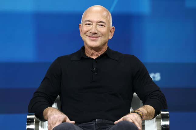 Jeff Bezos wearing a black long sleeve shirt and jeans smiling directly into the camera while sitting in a chair in front of a blue backdrop