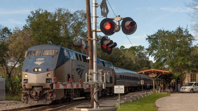 An Amtrak passenger train leaving DeLand Station , Florida at a level crossing with red lights.