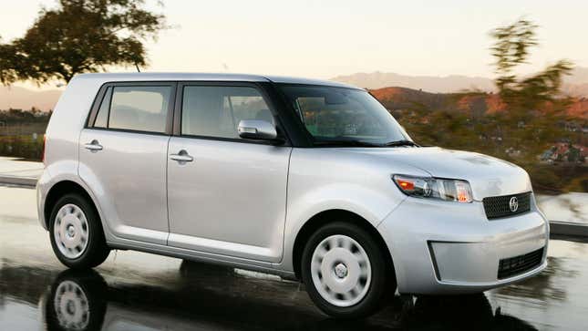 A photo of a silver Scion XB driving along a road. 