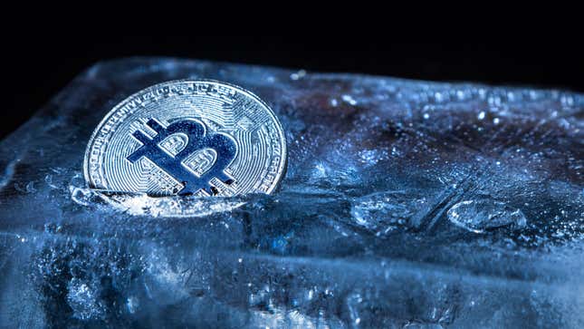 A gold bitcoin stuck partly in a block of ice, everything tinted blue.