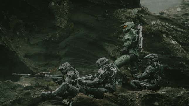 Three Spartan Silver team members sit on an outcropping of rocks while Master Chief stands next to them. 