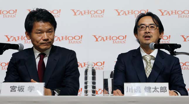 two men wearing suits and ties sitting behind microphones, yahoo japan logos on the backdrop behind them