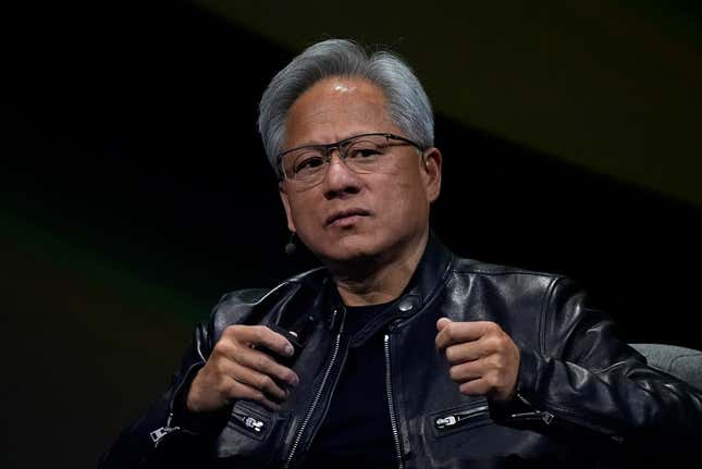 Jensen Huang wearing a black leather jacket sitting in a chair in front of a black backdrop with both his hands raised in almost fists