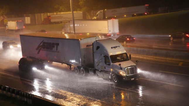 A photo of a semi truck driving near a small car on a wet highway. 