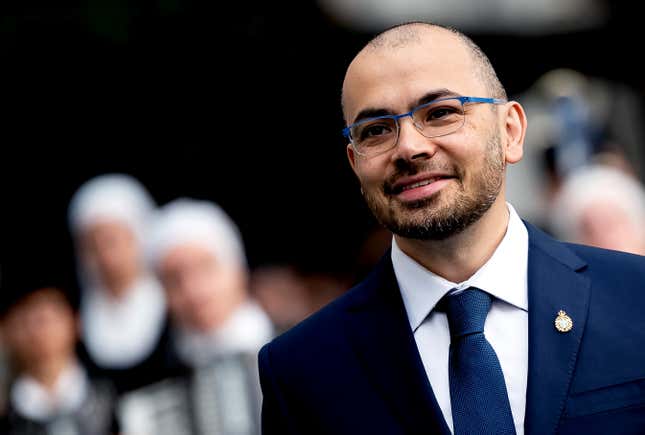 Demis Hassabis close up wearing a navy suit and tie, blurred people are behind him