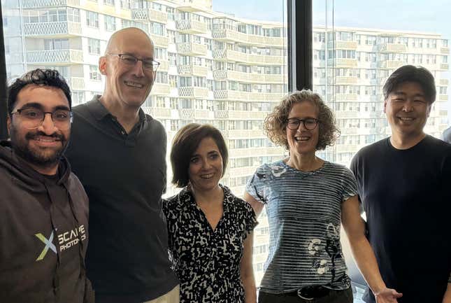 five people stand side by side, smiling, in front of a window with buildings in the background