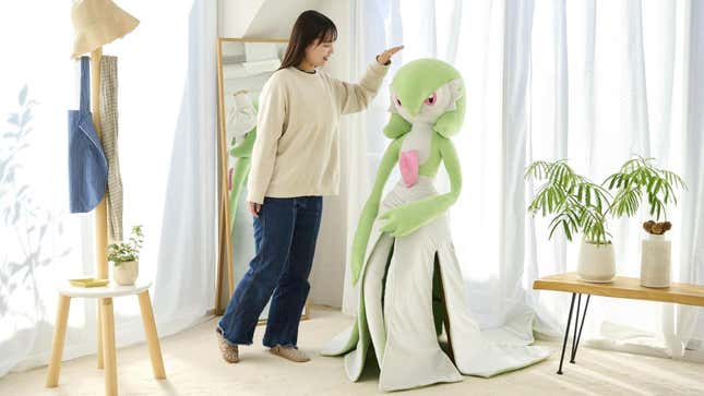 A person is shown standing next to the Gardevoir plush and patting it on the head.