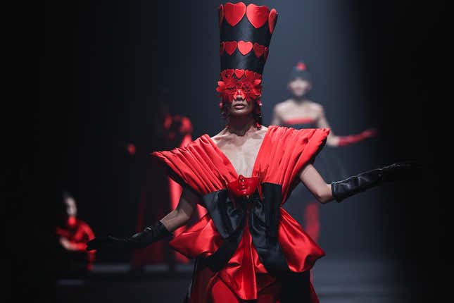 A woman walks down a runway in a dramatic red-and-black dress with a headpiece decorated with hearts extending over her eyes