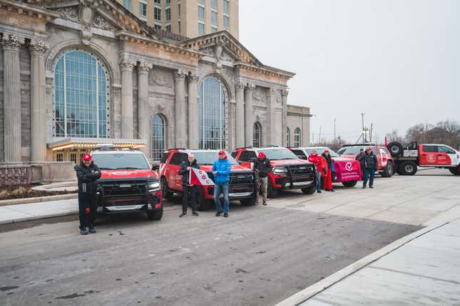 Five red and white F-150s line up in front of an old train station and pose for pictures, with several people standing outside the cars in winter gear holding flags.