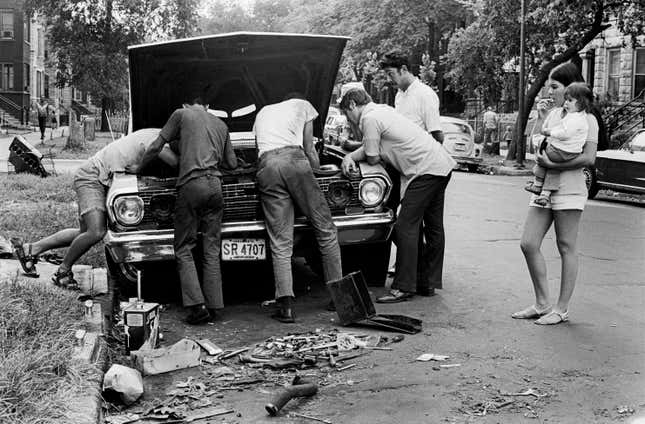 An old photo of a group of five men working under the hood of a car while a woman holding a baby watches