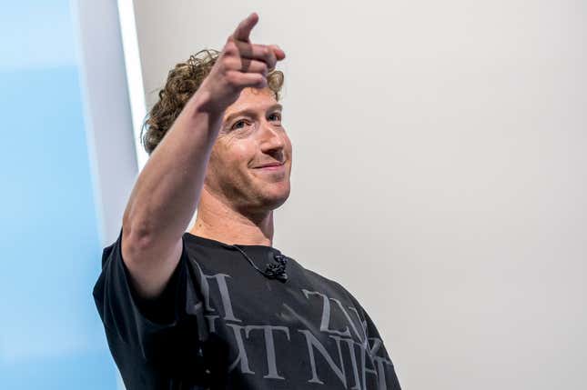 Mark Zuckerberg wearing a black t shirt with words printed on it, smiling and pointing with his right hand