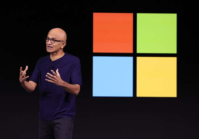 Satya Nadella speaking with his hands up in front of a black backdrop with a large Microsoft logo