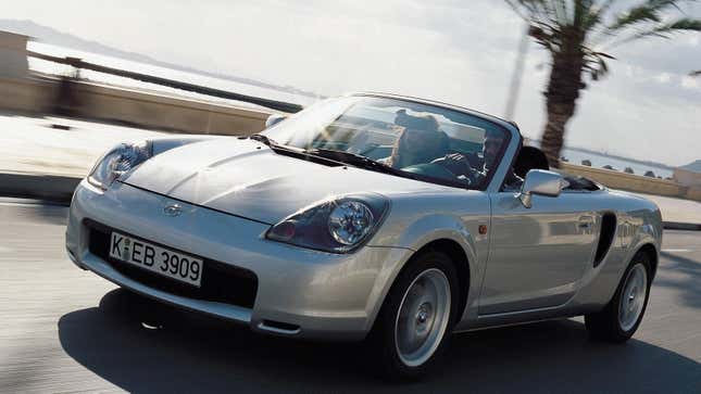 A silver Toyota MR2 Spyder driving next to a coastline with the top down