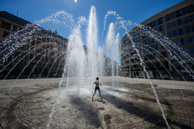 A kid runs through a large water fountain in the middle of a city street.