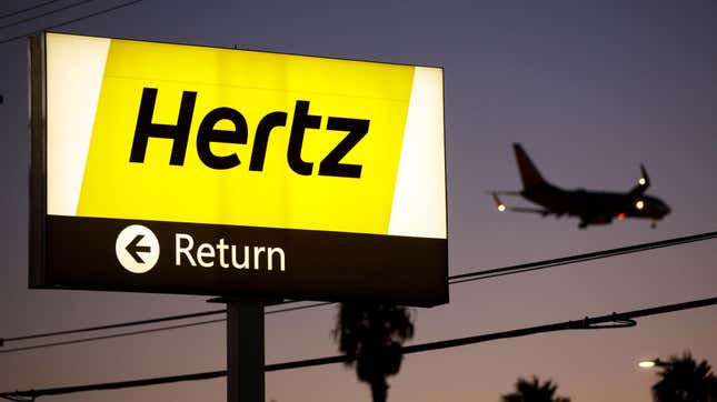 A Hertz rental car return sign at an airport with electrical wires and a plane in the background.