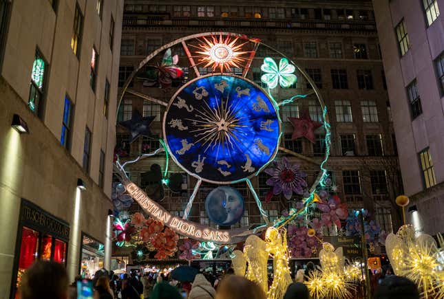 La multitud mira hacia una exhibición bien iluminada que muestra los signos del zodíaco y otros emblemas en el costado de un edificio de Saks