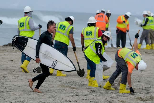 FILE - A surfer walks by as workers in protective suits continue to clean the contaminated beach in Huntington Beach, Calif., on Oct. 11, 2021. Federal regulators on Tuesday, Dec. 5, 2023, concluded the 2021 rupture of an undersea oil pipeline off the Southern California coast was likely caused by the proximity of anchored shipping vessels. (AP Photo/Ringo H.W. Chiu, File)