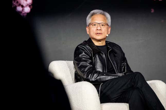 Jensen Huang wearing all black and a black leather jacket sitting in a white arm chair speaking with a lav mic on his face