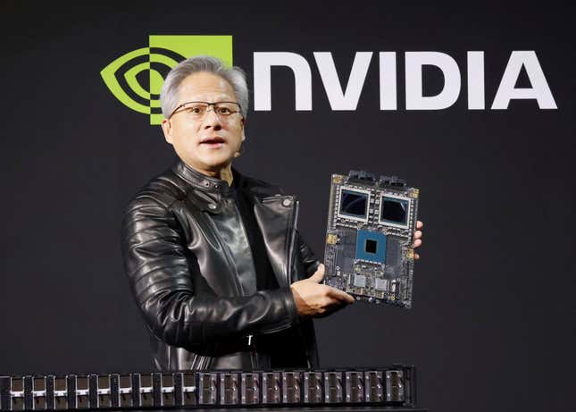 Jensen Huang wearing a black leather jacket holding up a chip board in front of a black backdrop with the Nvidia logo