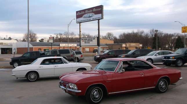 A red and a white Chevrolet Corvair share a parking lot with other modern cars.