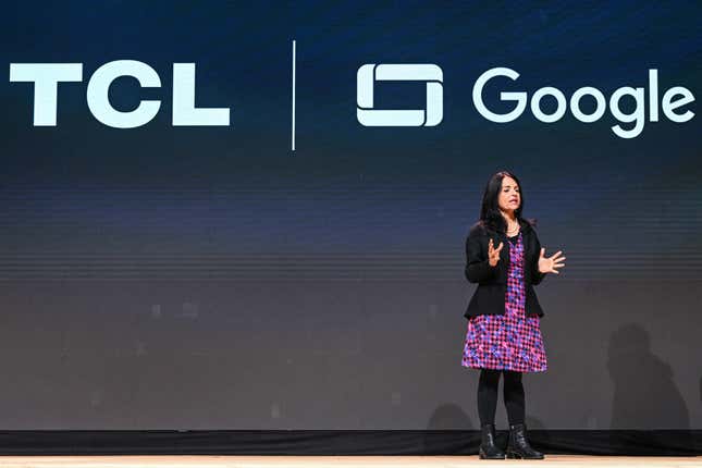 a woman wearing a purple patterned dress, black jacket, black tights and black boots on stage in front of a black backdrop that says TCL and Google