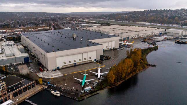 Boeing production facility in Renton, Washington: a warehouse with planes outside next to some trees and water with peopled hills in the background.