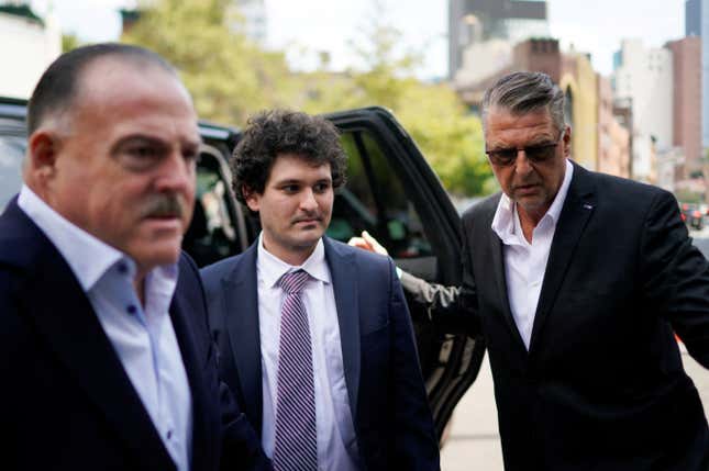 Sam Bankman-Fried, flanked by what appear to be two bodyguards in suits, steps out of a car on his way to court.