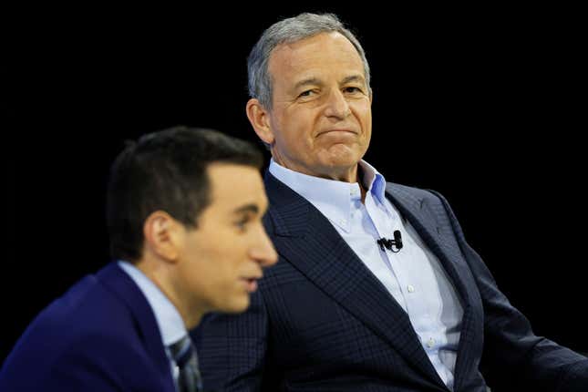 An in-focus Bob Iger (wearing a button down and suit jacket) looks to the left at an out-of-focus man