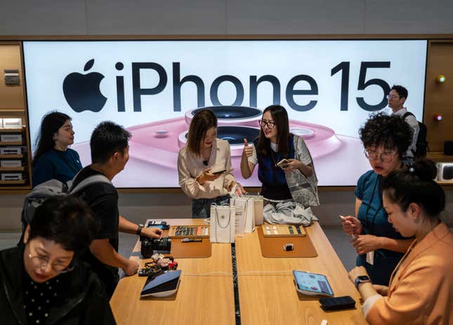 people surround a wooden table with Apple products displayed on it, an iPhone 15 sign is behind them