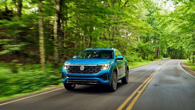 A blue Volkswagen Atlas Cross Sport driving down a heavily wooded road with bright green trees lining it