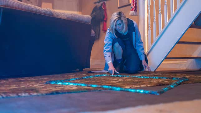 Jodie Whittaker's 13th Doctor examines a blue glowing square outline of energy on a living room's carpeted floor.