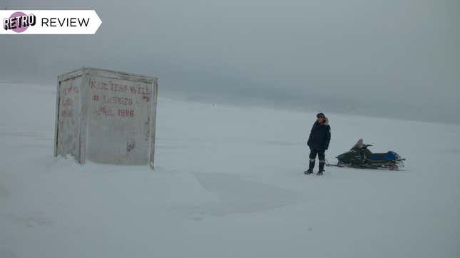 A man stands next to a small snowmobile on an icy landscape, staring at a box covering an oil test well.