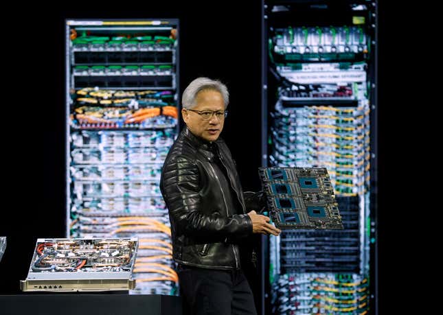 Jensen Huang wearing a black leather motorcycle jacket and fully black outfit holding a chip platform in front of a display of chip platforms in front of a backdrop with photos of server racks