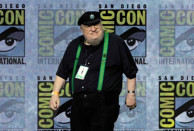 George R. R. Martin standing on a stage at Comic Con dressed in a black outfit with green suspenders.