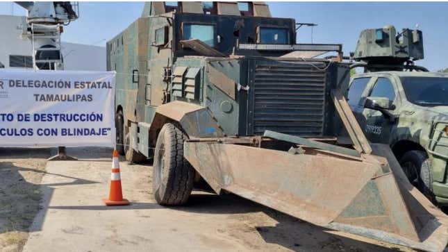 A large truck, seemingly military in nature, with a giant cow catcher on the front sits on a lot waiting for destruction. 