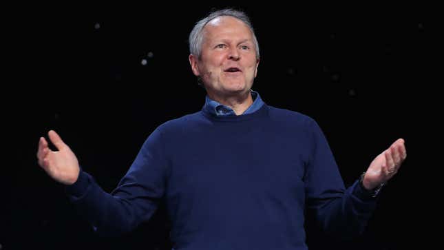 An aging man with thin, gray hair is standing happily against a dark backdrop.