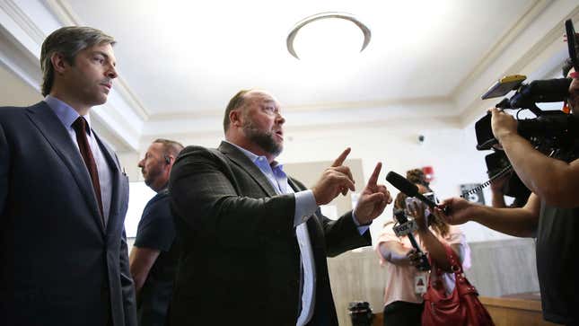 Alex jones stands in the center with both fingers up talking to reporters. His attorney Antonio Reynal stands behind him.