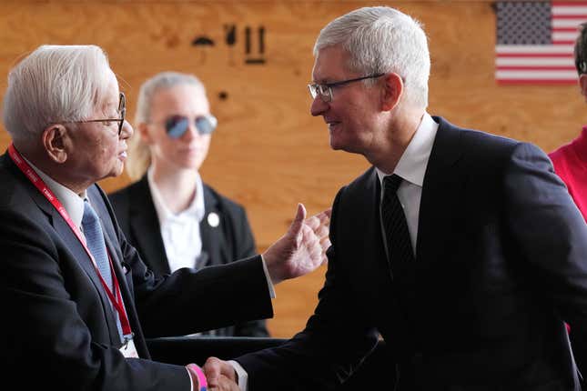 Morris Chang and Tim Cook shaking hands, a blonde secret service woman stands blurry behind them and a blurry American flag is in the top right corner