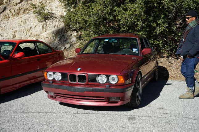 A red BMW E34 M5 is parked with a fashionable man standing next to it.