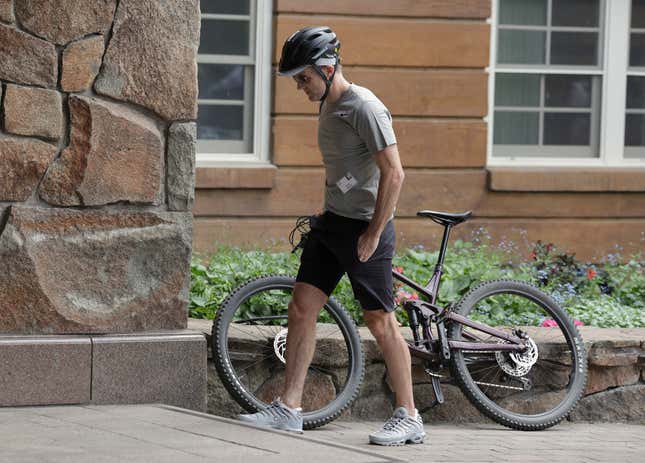 a man in a grey t shirt, black shorts, and grey shoes walking beside his bike in front of a wood and stone building