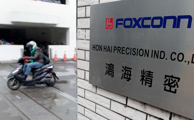 A close up of a silver placard sign readings "Foxconn" and underneath "Hon Hai Precision Ind." on the outside of a white-tiled building. A motorcyclist drives past in the background.