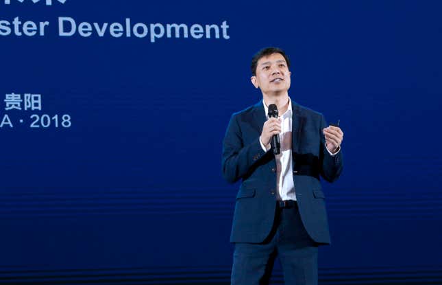 Robin Li speaking into a microphone wearing a navy blue suit jacket and pants in front of a dark blue backdrop