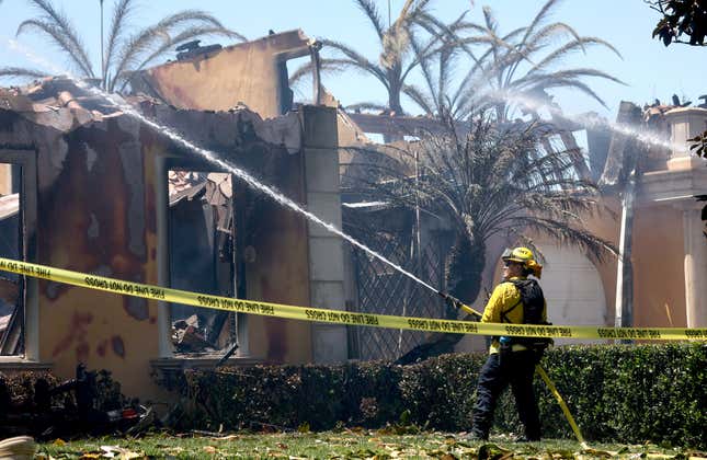 A firefighter aims a hose at a fire in front of a destroyed home. A yellow "fire line do not cross" tape cuts across the image.