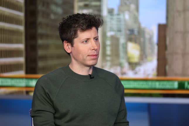 Sam Altman wearing a greenish grey sweatshirt with a small smile on his face as he looks at someone he's talking to